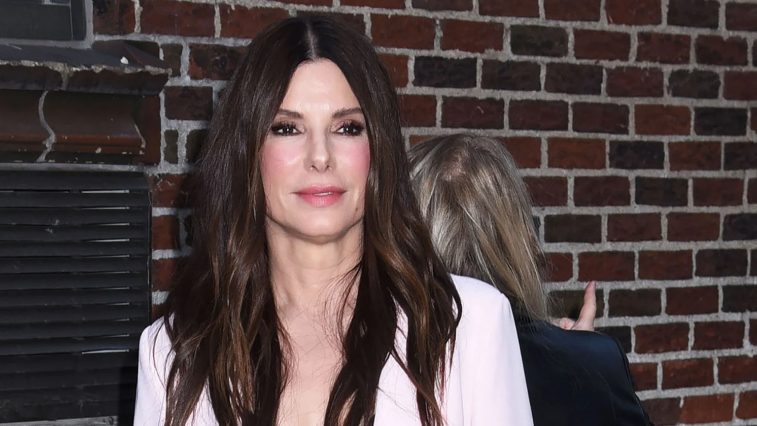Sandra Bullock with long, wavy dark brown hair and a pink top, smiling softly in front of a brick wall.