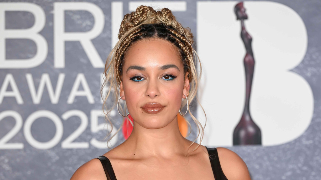 Jorja Smith poses at the BRIT Awards 2025, wearing blue eyeliner and glossy lips, with her blonde braids styled in two buns.