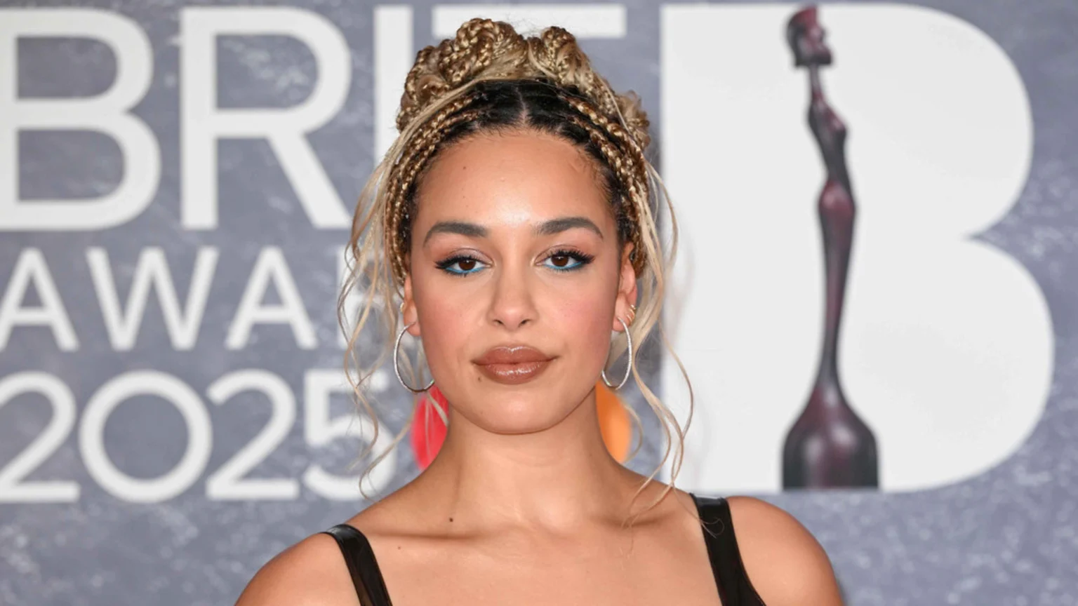 Jorja Smith poses at the BRIT Awards 2025, wearing blue eyeliner and glossy lips, with her blonde braids styled in two buns.
