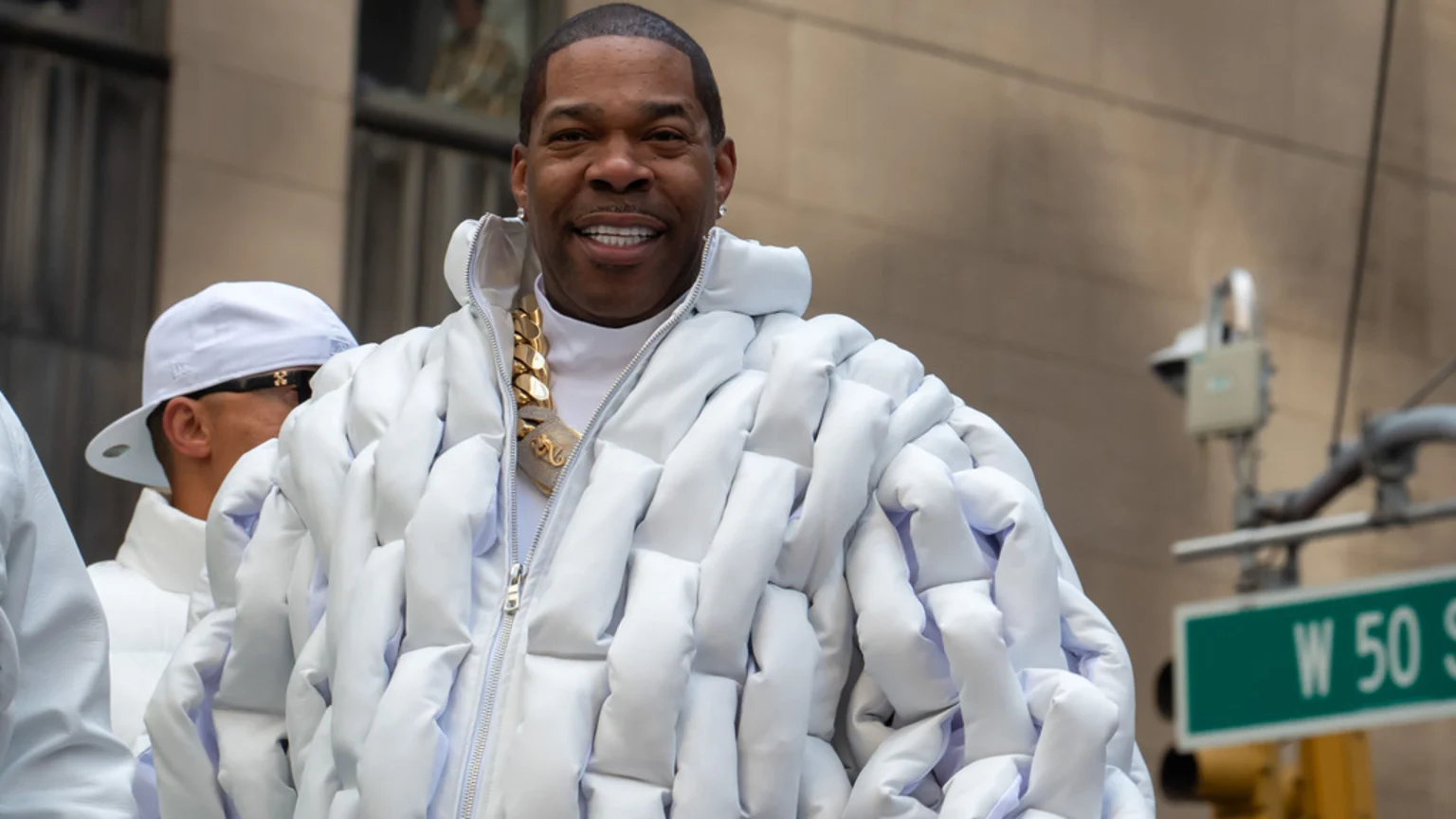 A smiling man with a large gold chain and a unique white woven puffer jacket stands on a street with a "W 50 ST" sign.