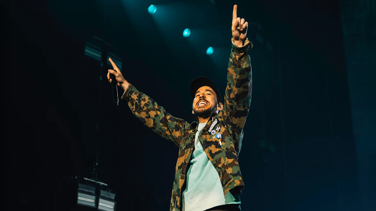 A smiling performer in a camouflage jacket and baseball cap holds a microphone in one hand and points skyward with the other, on a dark stage with teal lights.