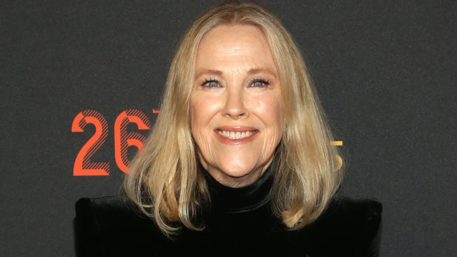 A close-up studio portrait of Candice Bergen smiling broadly. She wears a black high-neck velvet-like top, with shoulder-length blonde hair and blue eyes, illuminated by bright, even lighting. The dark background features large orange numbers with a diagonal stripe pattern.