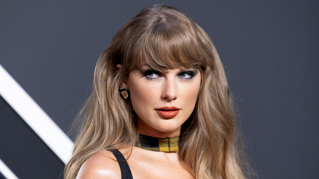 Close-up of Taylor Swift with long blonde hair and bangs, dark eye makeup, and orange-brown lipstick. She wears a plaid choker and a yellow earring against a dark gray background.