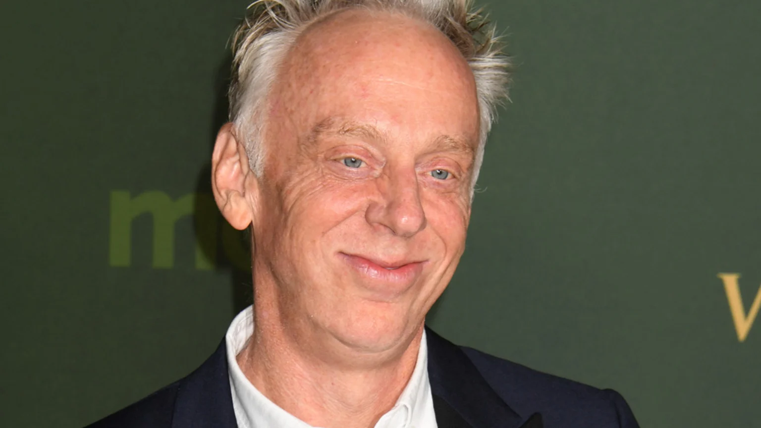 A smiling man with spiky gray hair and blue eyes wears a white shirt and dark suit jacket against a dark green background with a faint logo.