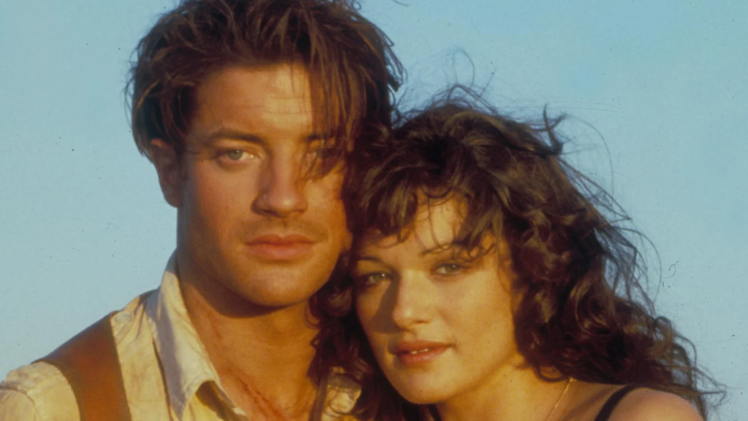 A close-up of actors Brendan Fraser and Rachel Weisz posing together against a light blue sky. Fraser wears a light shirt and suspenders, and Weisz has dark, curly hair and wears a dark strap top.