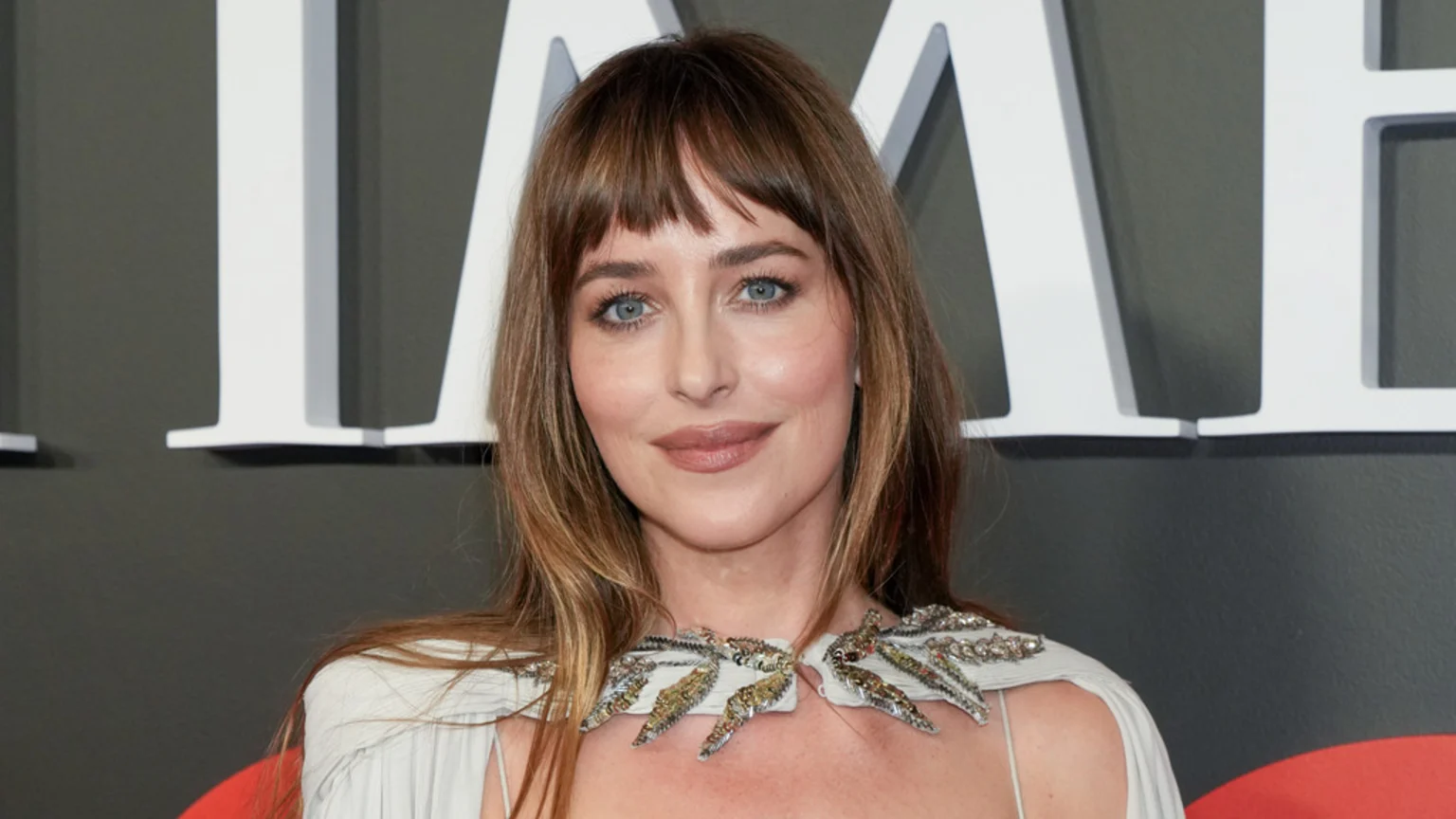 Dakota Johnson with long brown hair and bangs, wearing a light dress with embellished shoulders, smiles directly at the camera against a dark background with large white letters.