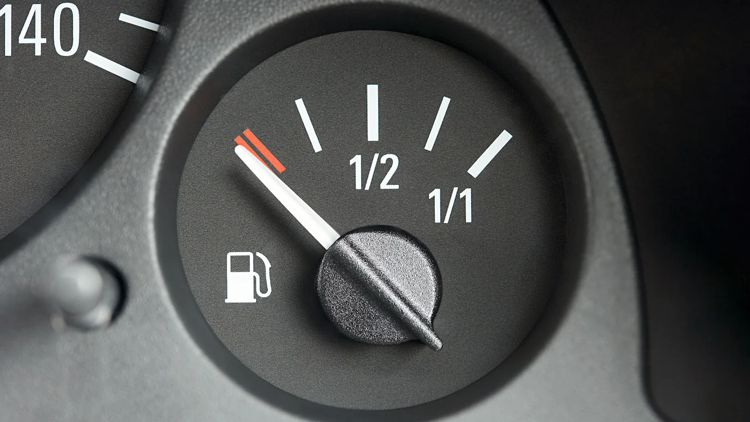 A car's fuel gauge shows the needle pointing to the red, near-empty section next to a fuel pump icon, with '1/2' and '1/1' markings visible.