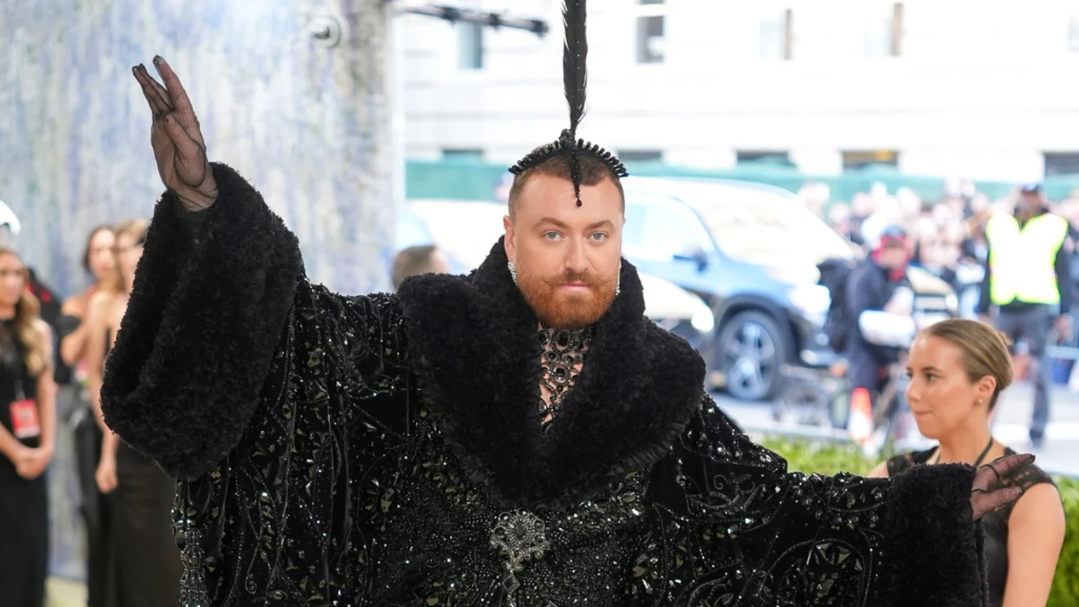Sam Smith, with a reddish beard and mustache, wears an elaborate black beaded coat with a furry collar and a feather headpiece, raising one arm in a sheer black glove at an event.