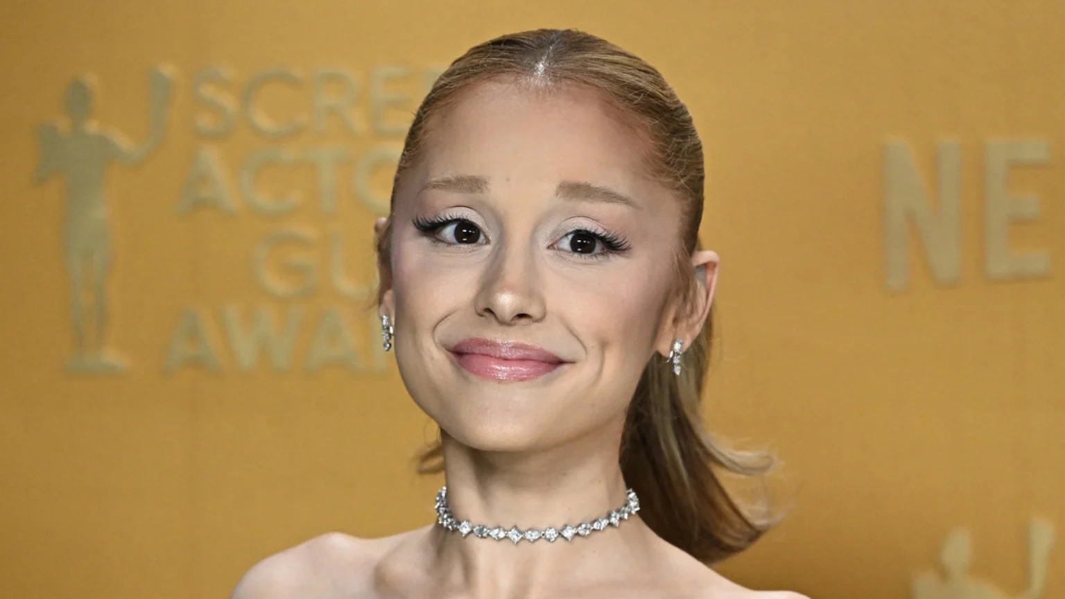 Ariana Grande smiles at the camera, wearing a diamond choker and earrings, with a blurred "Screen Actors Globe Awards" background.