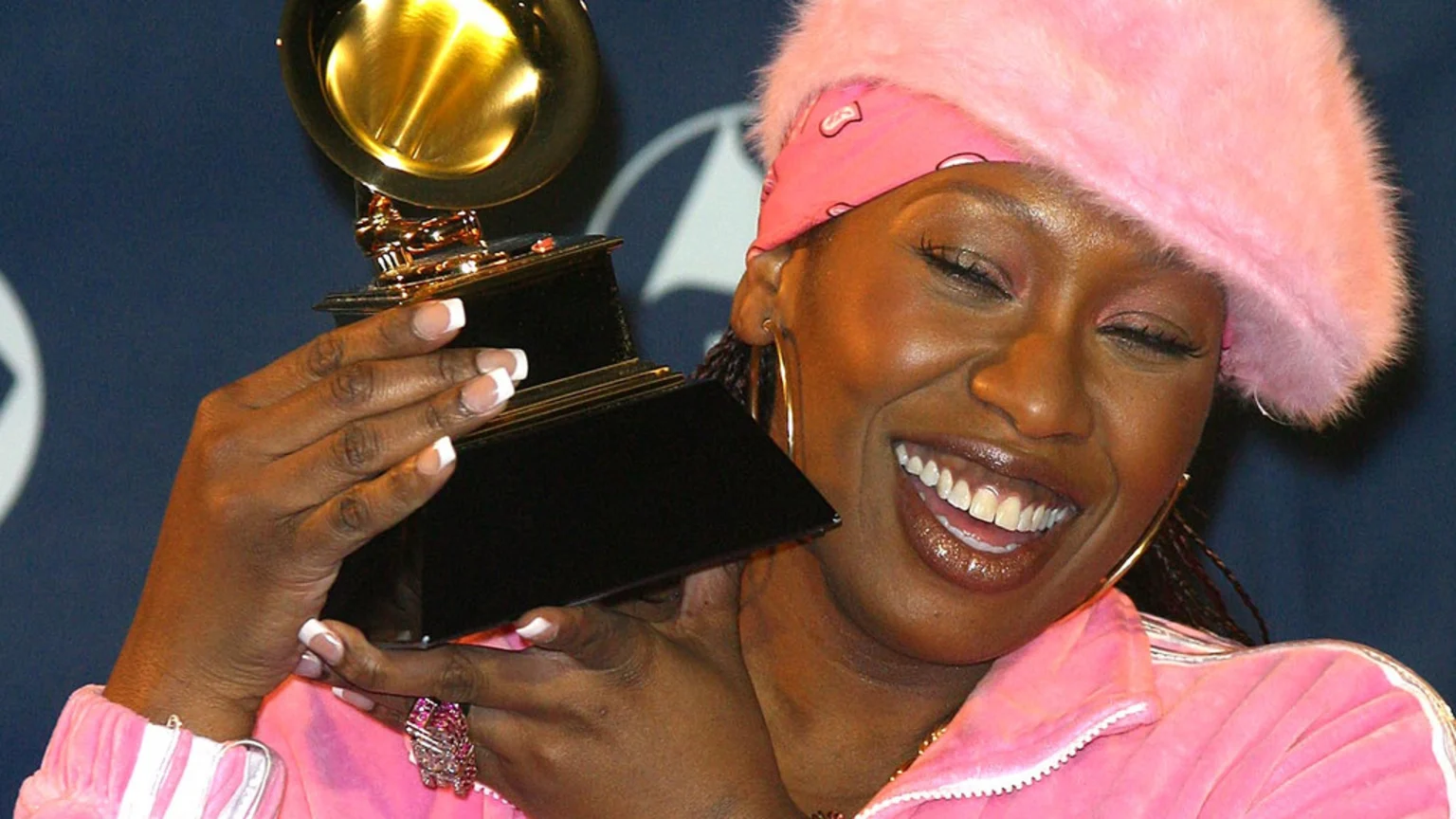 A joyful person with a pink fuzzy hat and tracksuit smiles with closed eyes while holding a golden Grammy award.