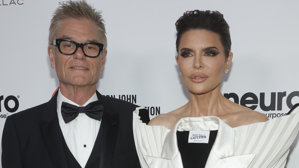 Harry Hamlin, in a black tuxedo and glasses, stands next to Lisa Rinna, who wears a white and black strapless top with dramatic makeup, against a white background.