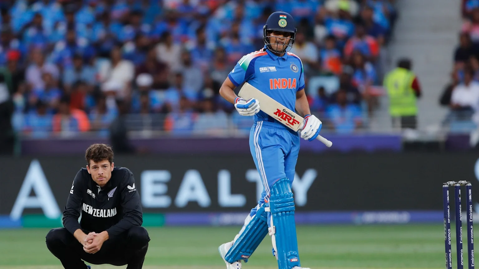 An Indian cricketer in blue uniform walks with a bat past a squatting New Zealand cricketer in black, with cricket stumps visible on the right.