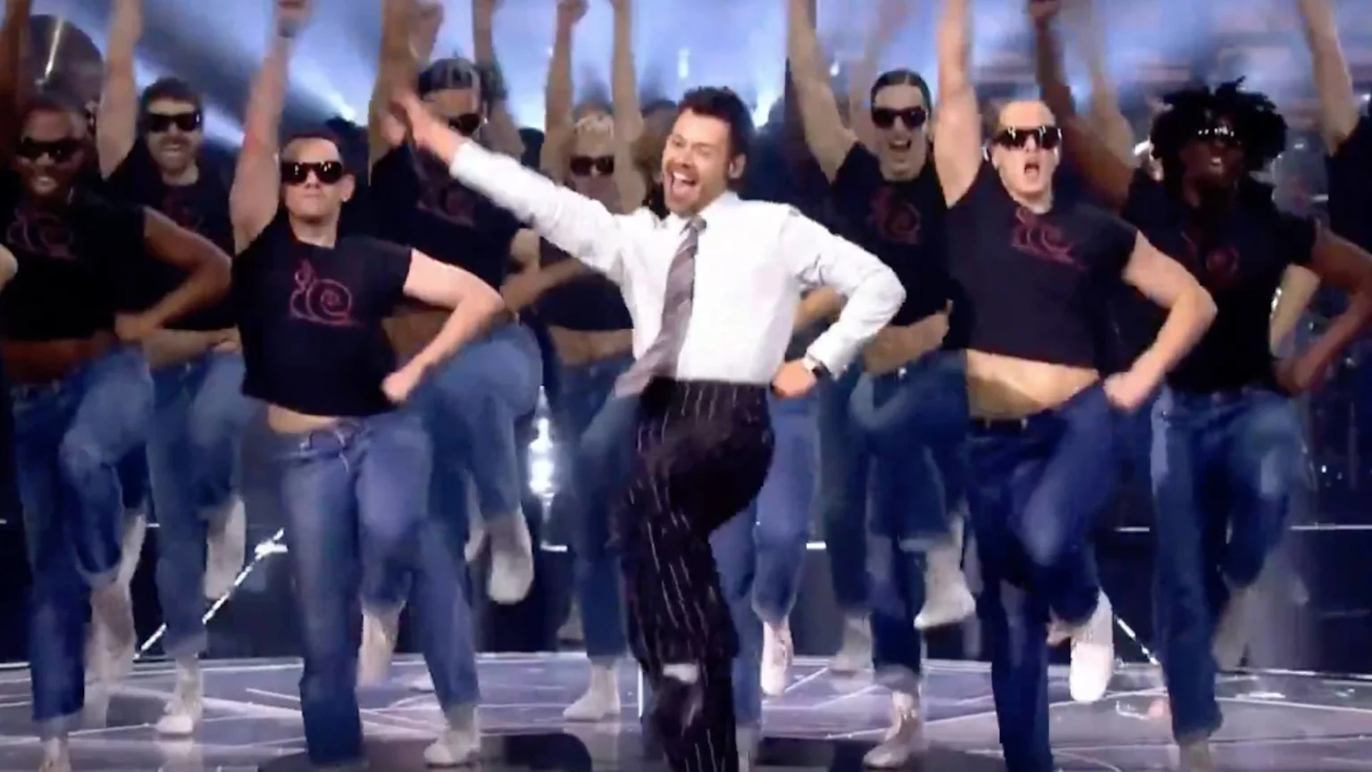 Harry Styles, in a white shirt and pinstripe pants, enthusiastically dances on stage with a wide smile, surrounded by a group of backup dancers in sunglasses, black tops, and jeans.