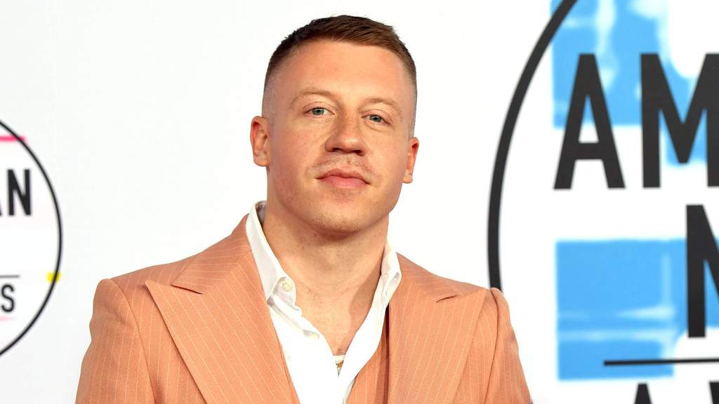Macklemore, a man with short brown hair, wearing a peach pinstripe suit and white shirt, looks forward in front of a white backdrop with partially visible "American Music Awards" logos.