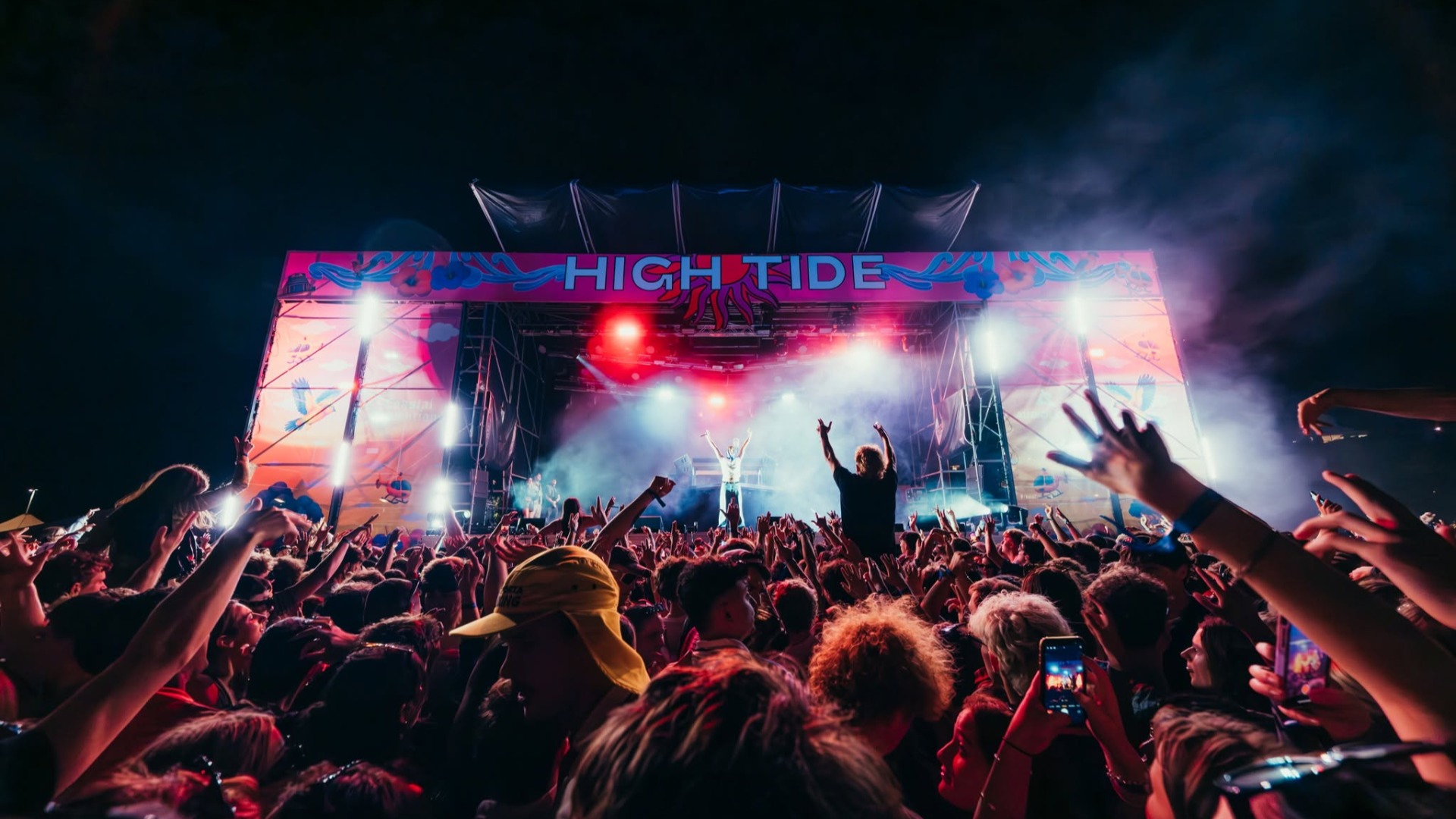 A vibrant nighttime music festival scene with a large crowd cheering, hands raised towards a brightly lit stage featuring performers and a "HIGH TIDE" banner.