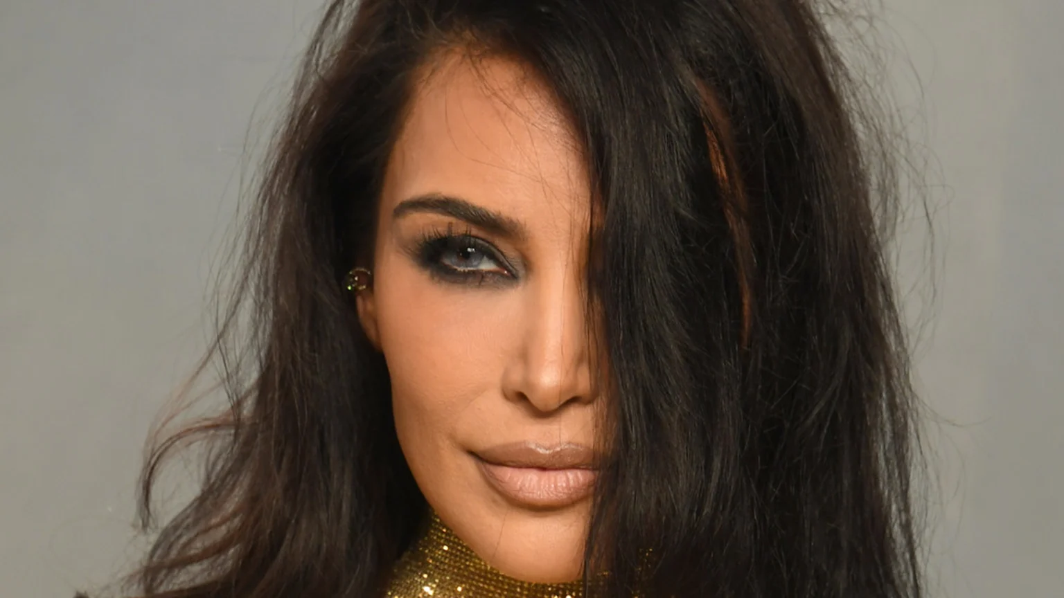 A close-up portrait of a woman with long dark hair covering part of her face, dramatic smokey eye makeup, light lips, and a gold beaded collar.