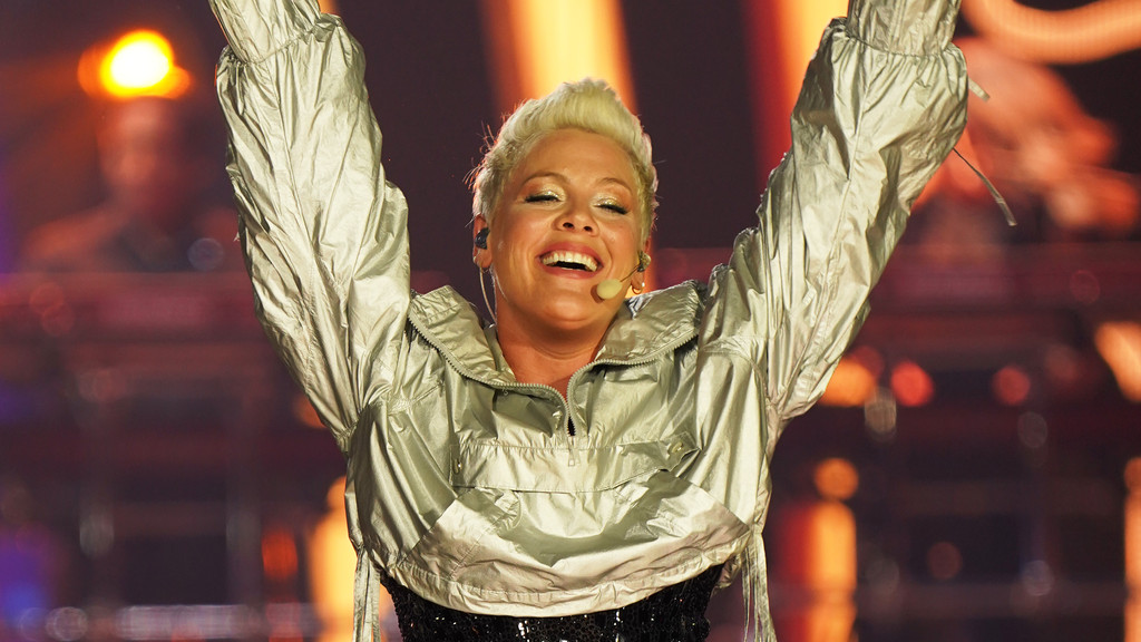 A woman with short, spiky, light pink-blonde hair and glittery eye makeup smiles widely, arms raised, wearing a shiny silver jacket and a black sequined top against a blurry stage background with orange lights.