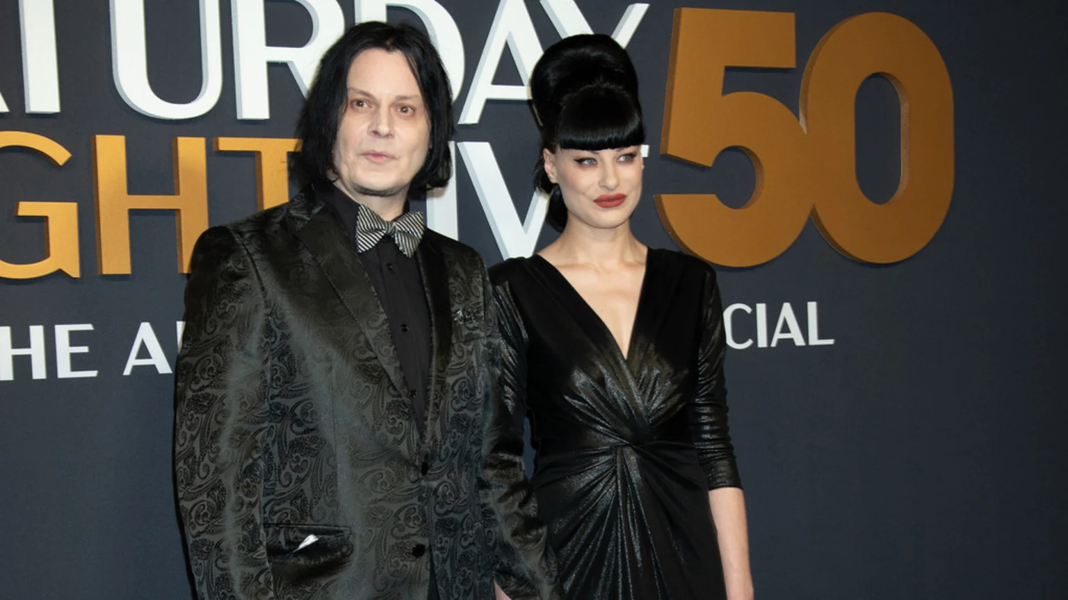 Jack White and Olivia Jean stand together at an event, with White in a patterned suit and Jean in a shiny black dress, in front of a "Saturday Night Live 50" sign.