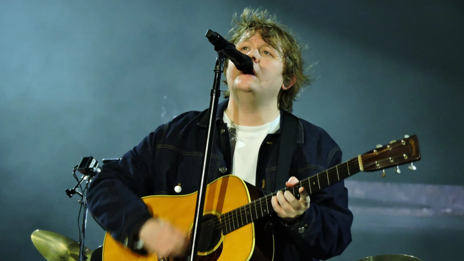 Lewis Capaldi, with messy brown hair, sings into a microphone while playing an acoustic guitar on a dark, smoky stage.