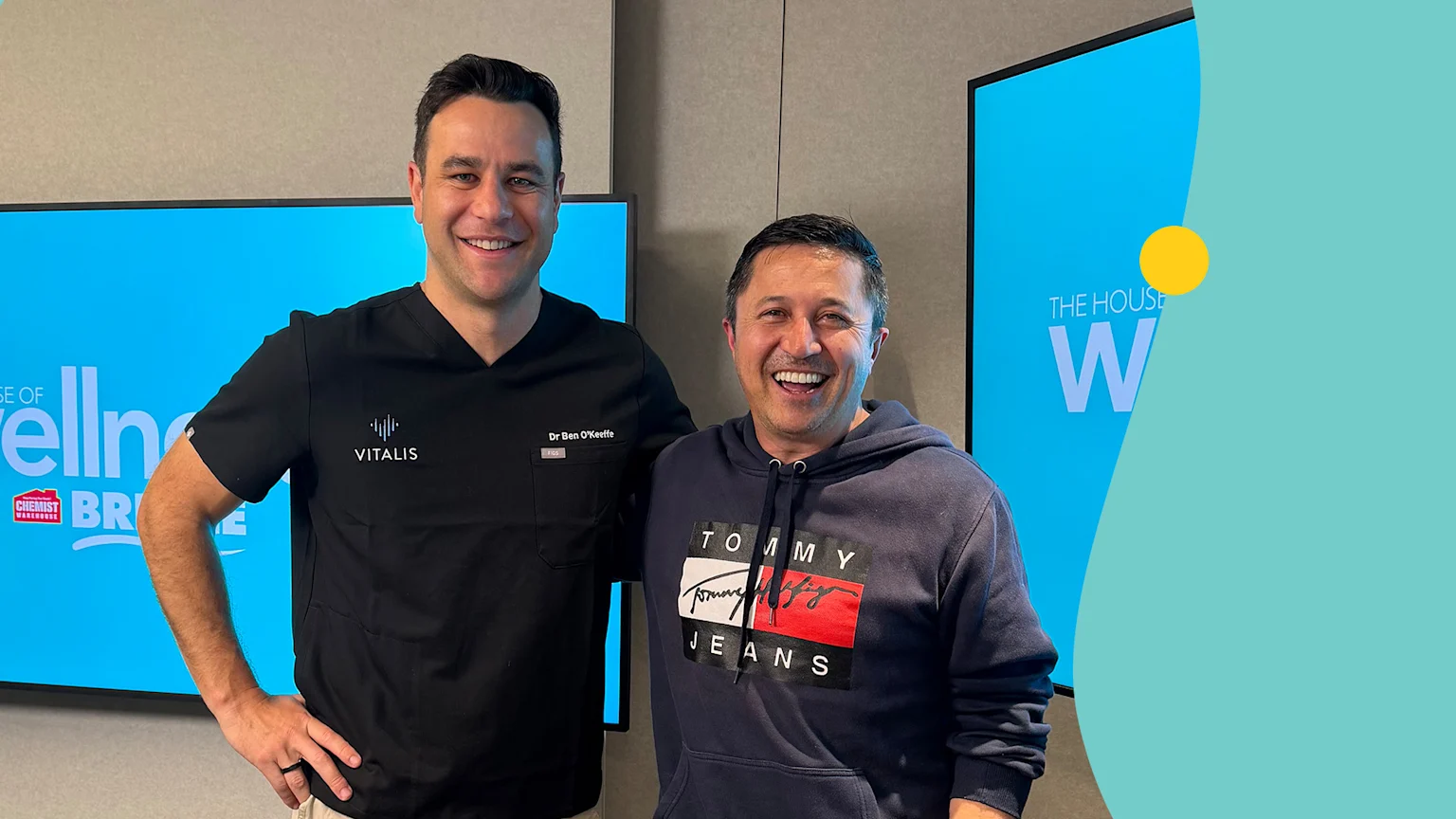 Dr. Ben O'Keeffe, in a black scrub top, stands smiling with another man in a navy hoodie, in front of screens showing 'THE HOUSE OF Wellness' branding.