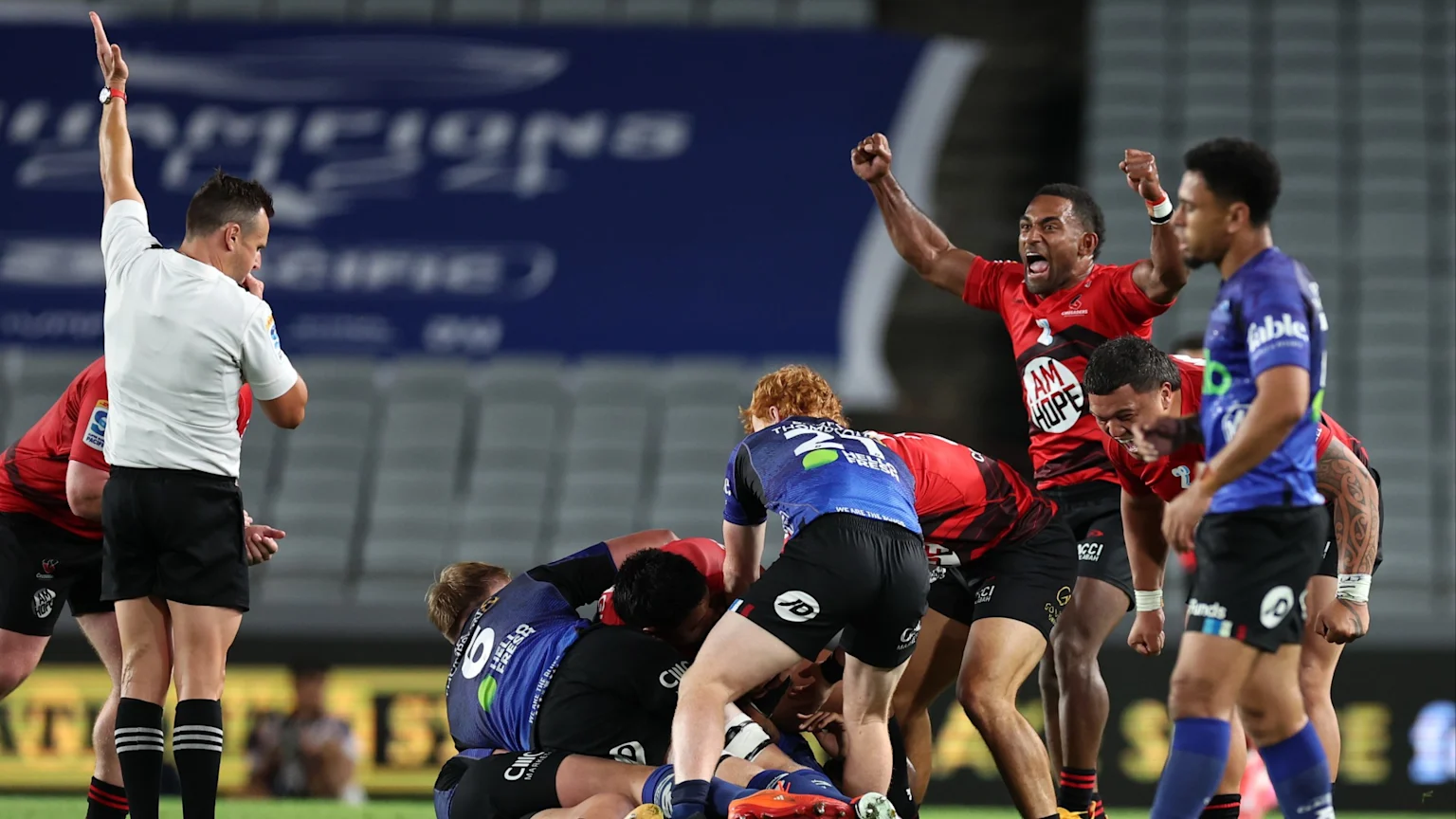A rugby referee signals during a match while players from two teams, one in red and one in blue, are engaged in a ruck, and red-jerseyed players celebrate.