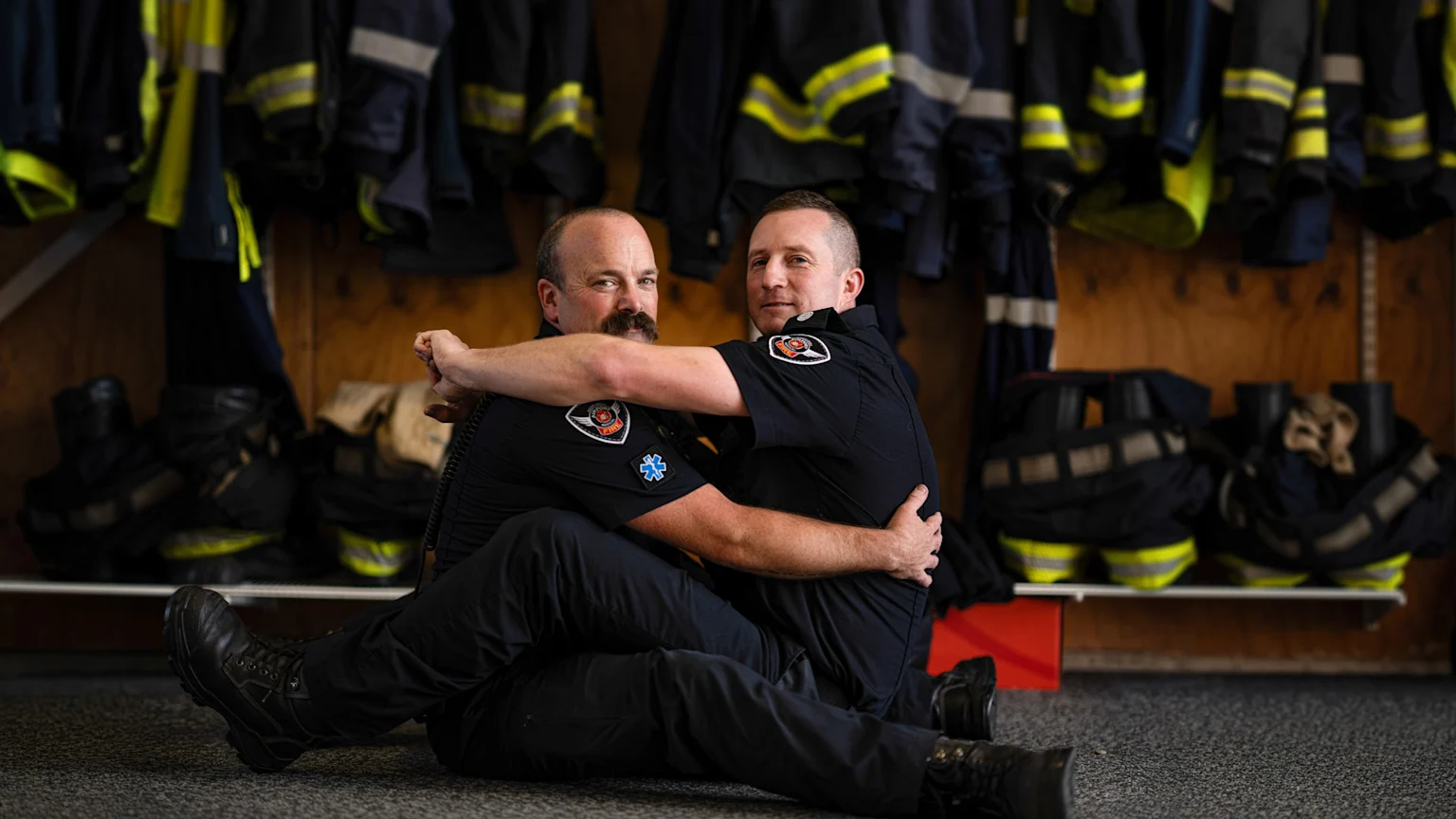 Two men in dark firefighter uniforms sit on the floor, embracing and smiling at the camera, with fire gear on shelves in the background.