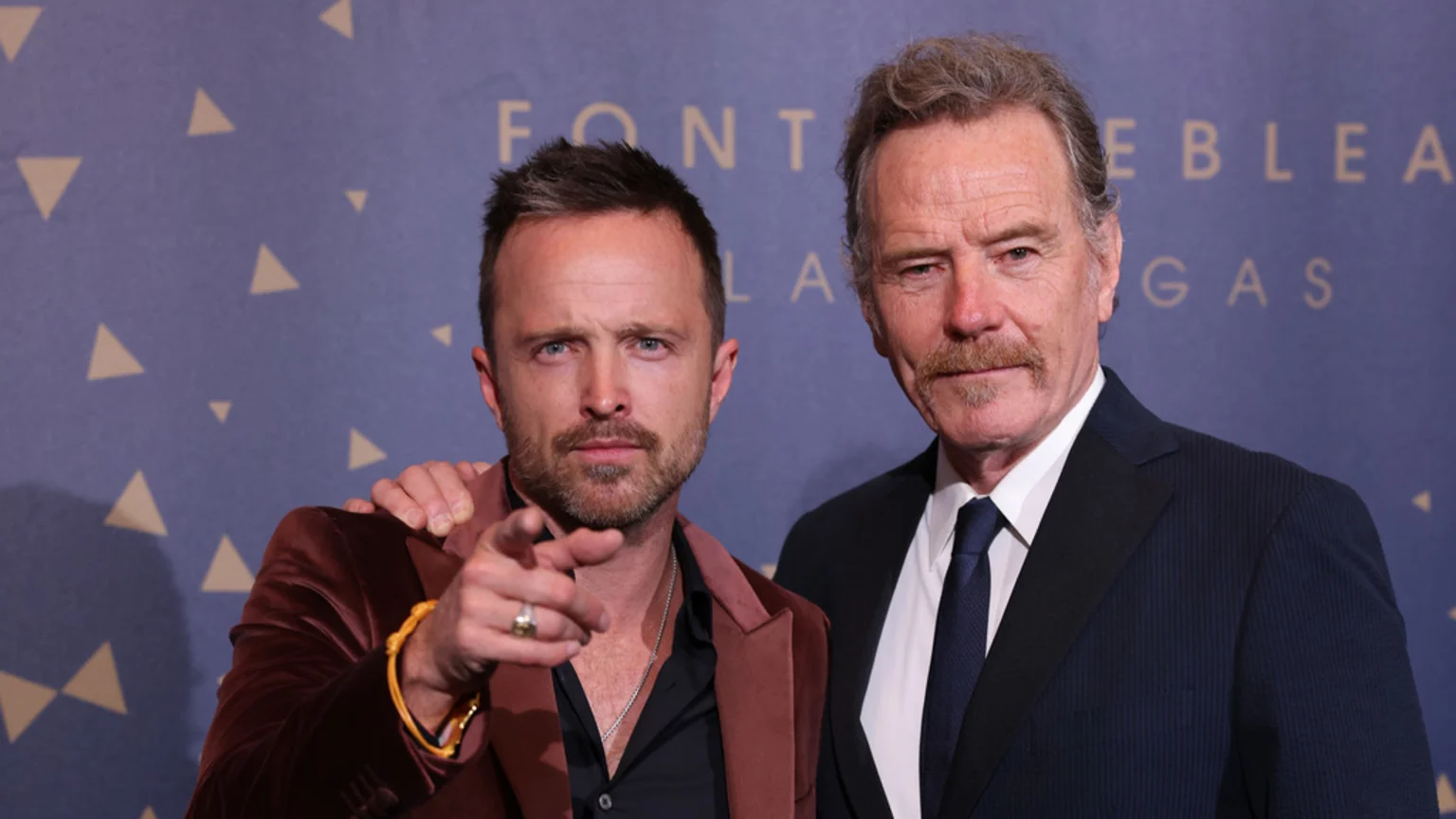 Bryan Cranston in a dark suit and Aaron Paul in a velvet blazer pose together, Paul pointing forward with his arm around Cranston, against a blue patterned backdrop.