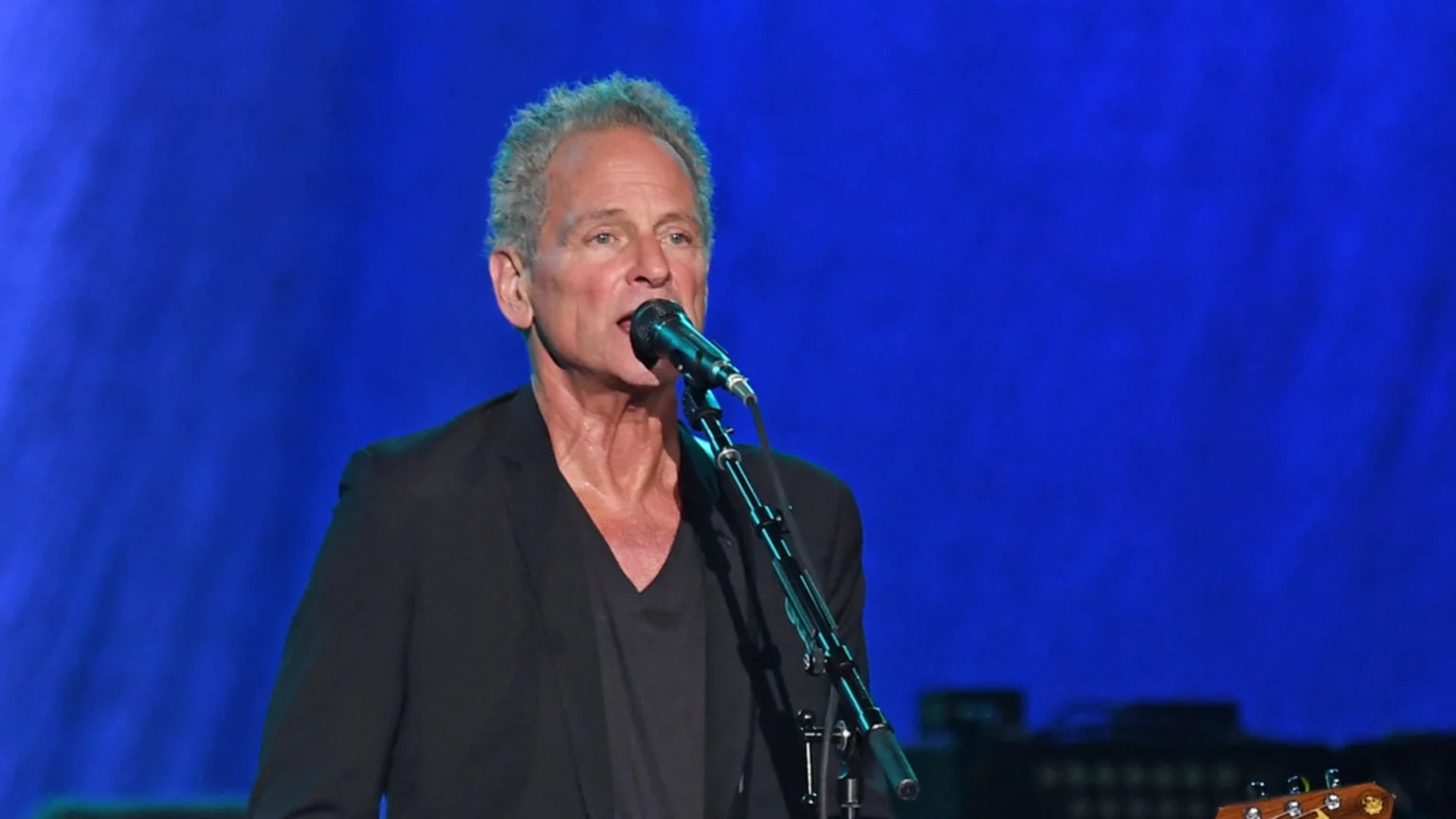 Lindsey Buckingham, a man with gray hair and a black shirt, sings into a microphone during a live performance against a vibrant blue background.