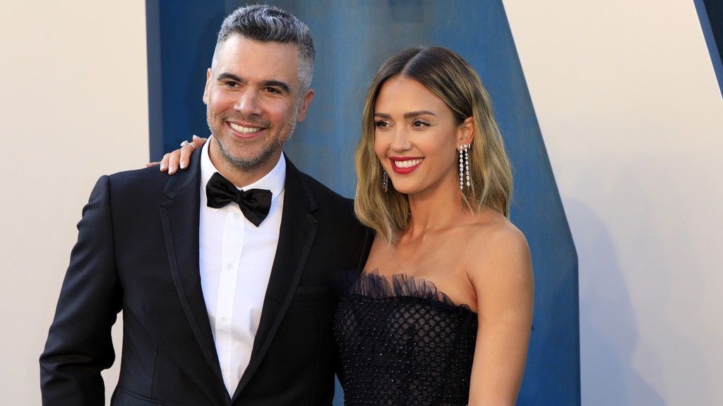Jessica Alba and Cash Warren smiling broadly for the camera. Alba wears a black strapless gown with textured details and long diamond earrings, with her hand resting on Warren's shoulder. Warren is in a classic black tuxedo with a bow tie. They are against a light-colored wall with a prominent blue diagonal section.