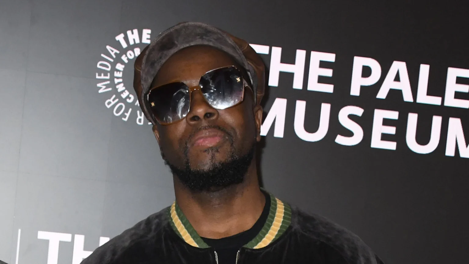Wyclef Jean wearing sunglasses, a dark cap, and a dark jacket with striped trim, standing in front of a sign for "The Paley Center for Media."