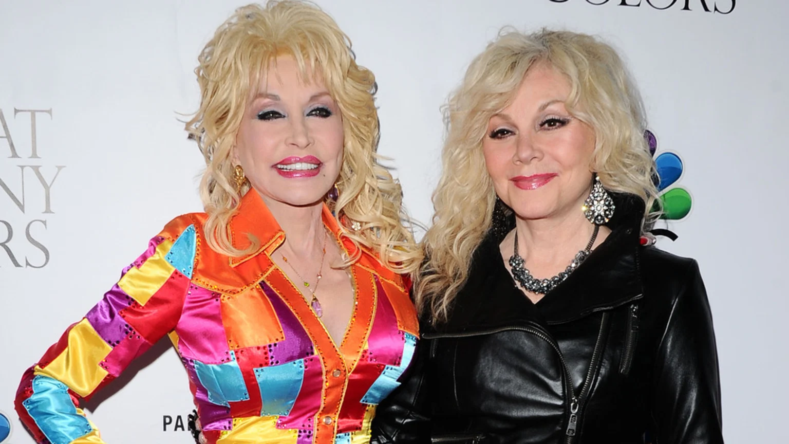 Dolly Parton in a colorful patchwork top and Stella Parton in a black leather jacket smile, standing together against a white background with a logo.