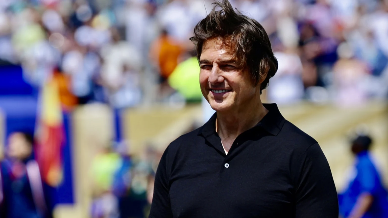Tom Cruise, smiling in a black polo shirt, stands outdoors with a blurred crowd and colorful flags in the sunlit background.