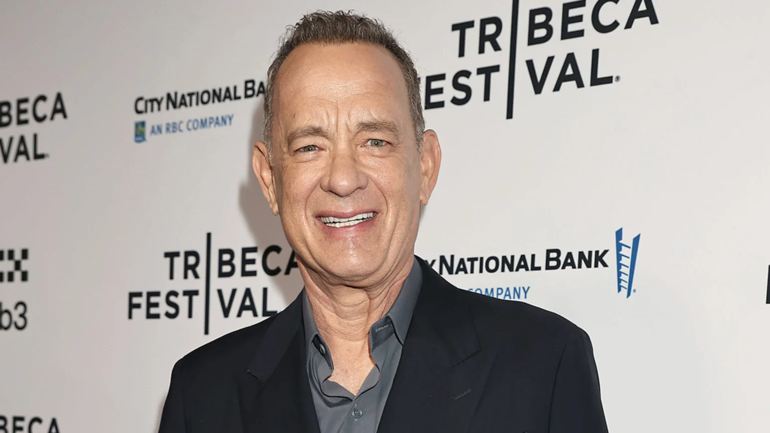 Tom Hanks smiles at the camera, wearing a dark suit jacket and grey shirt, in front of a white backdrop with "Tribeca Festival" and "City National Bank" logos.
