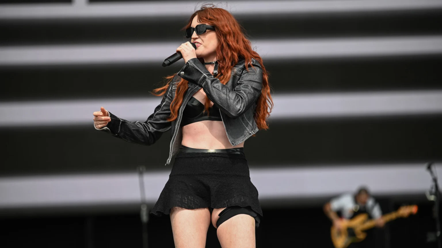A woman with long red hair and sunglasses sings into a microphone on stage, wearing a black leather crop jacket, bra, and mini-skirt, with a blurry guitarist in the background.