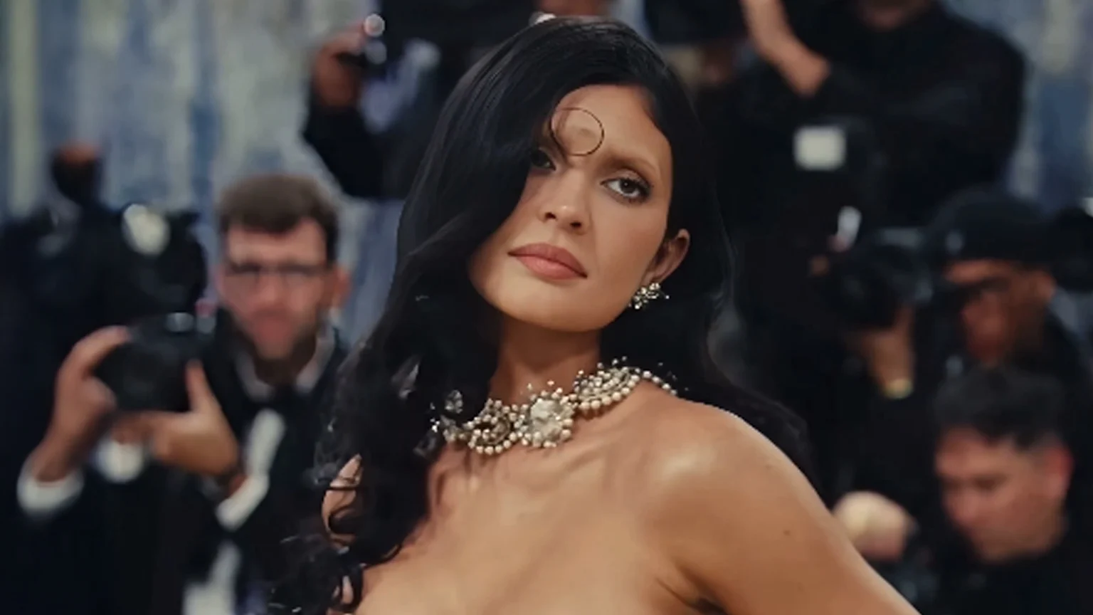 Kylie Jenner, with dark hair, light eyebrows, and a circular forehead design, wears a large pearl necklace on bare skin and looks at the viewer amidst a blurred crowd of photographers.