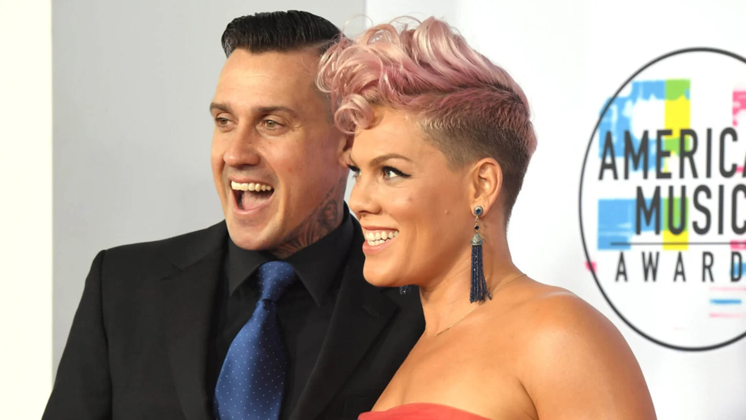 A man in a black suit and a woman with pink hair and a strapless top are smiling broadly, standing in front of an "American Music Awards" logo.