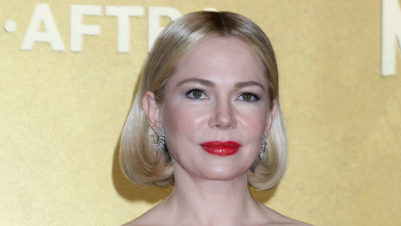 Michelle Williams poses with blonde bob hair, red lipstick, and sparkling earrings against a golden-yellow background with blurred white text.