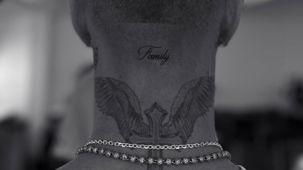 A close-up grayscale photograph of the back of an adult's neck showing 'Family' tattoo in script font, a winged cross tattoo, and two silver necklaces—a link chain and a beaded chain with decorative spheres. The background is blurred indoors with shallow depth of field.