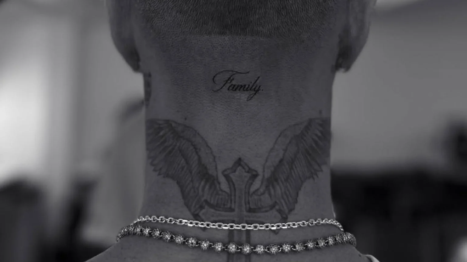 A close-up grayscale photograph of the back of an adult's neck showing 'Family' tattoo in script font, a winged cross tattoo, and two silver necklaces—a link chain and a beaded chain with decorative spheres. The background is blurred indoors with shallow depth of field.