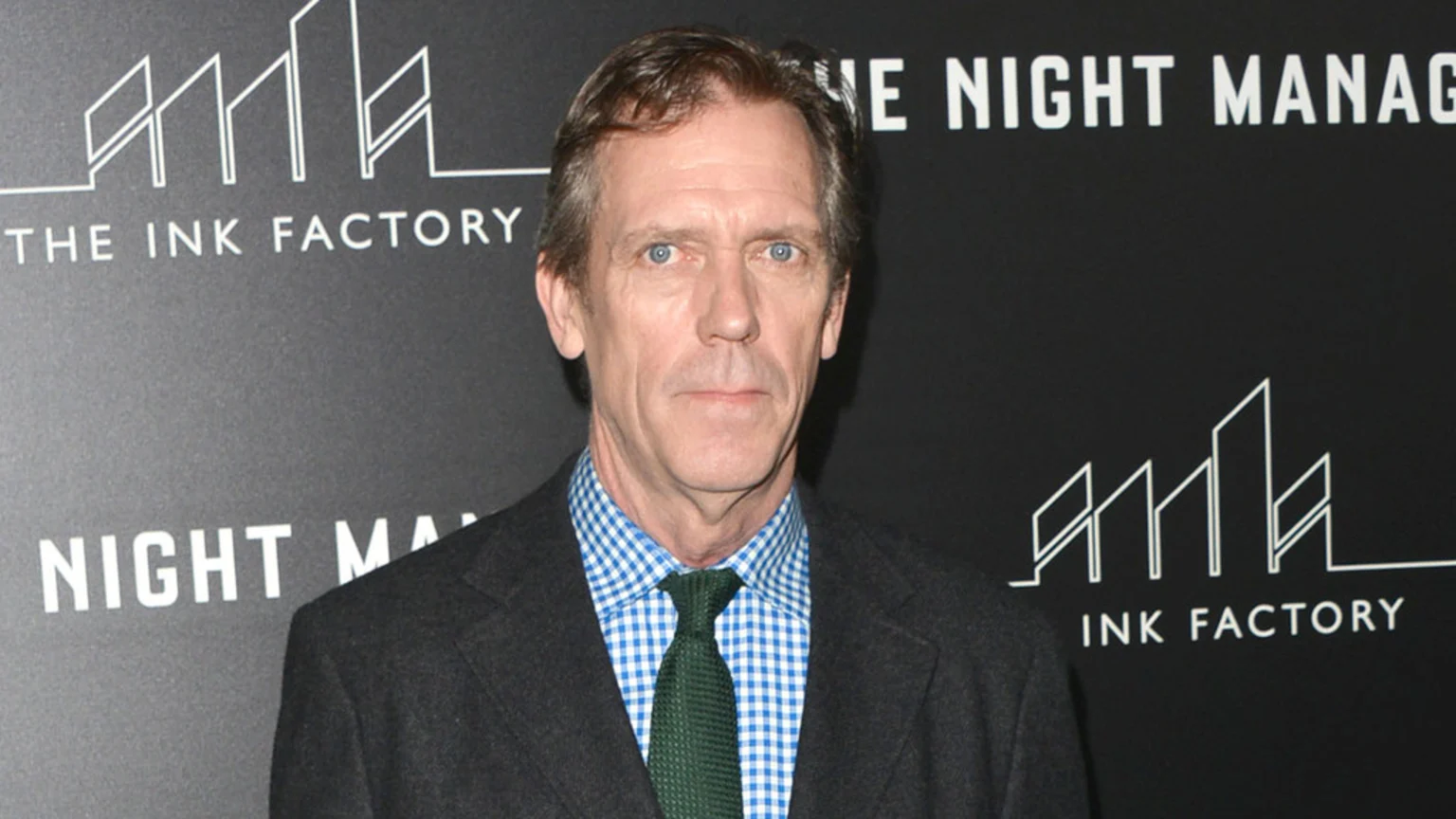A headshot of British actor Hugh Laurie, a middle-aged man with brown hair and blue eyes, looking directly at the camera with a neutral expression. He wears a blue and white gingham patterned shirt, a dark green knitted tie, and a charcoal blazer. Behind him is a dark grey backdrop with the white text "THE INK FACTORY" and "THE NIGHT MANAGER" partially visible, along with a stylized skyline logo.