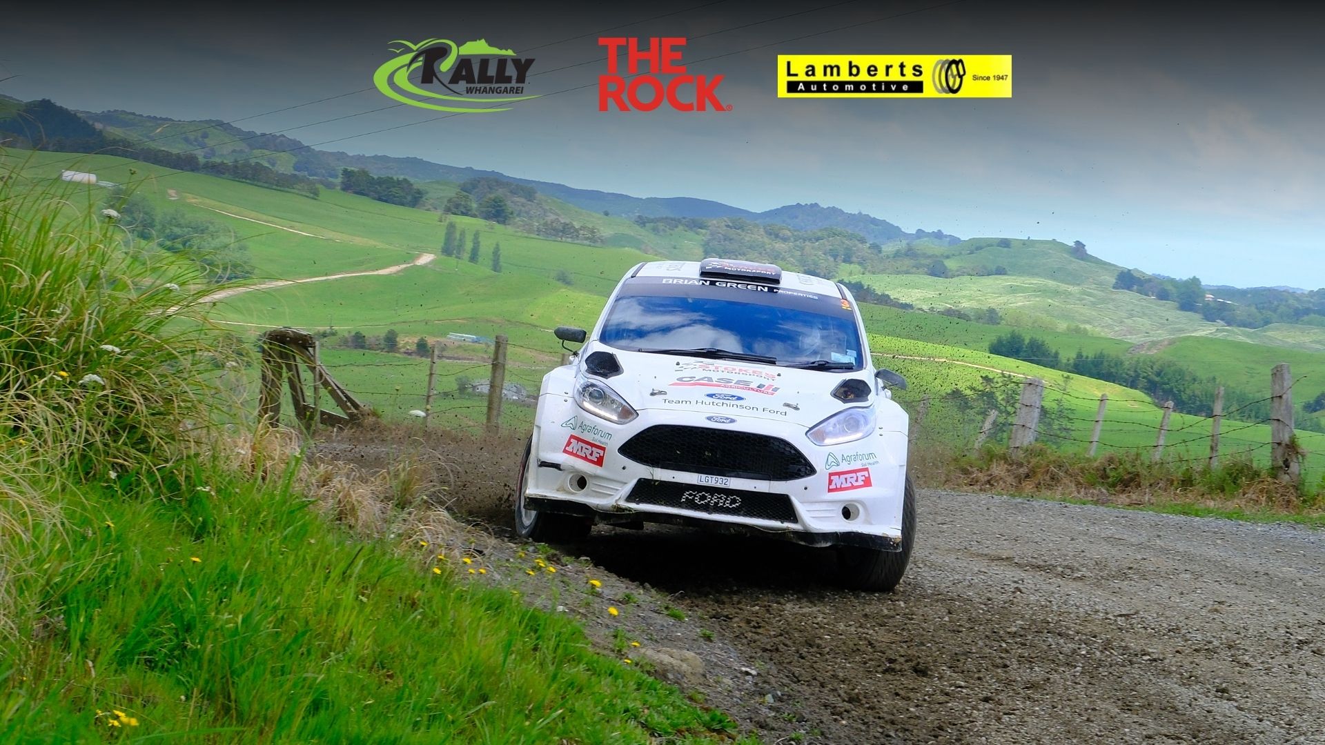 A white rally car speeds along a dirt road, kicking up dust, with green rolling hills, a fence, and event sponsor logos visible in the background and top of the image.