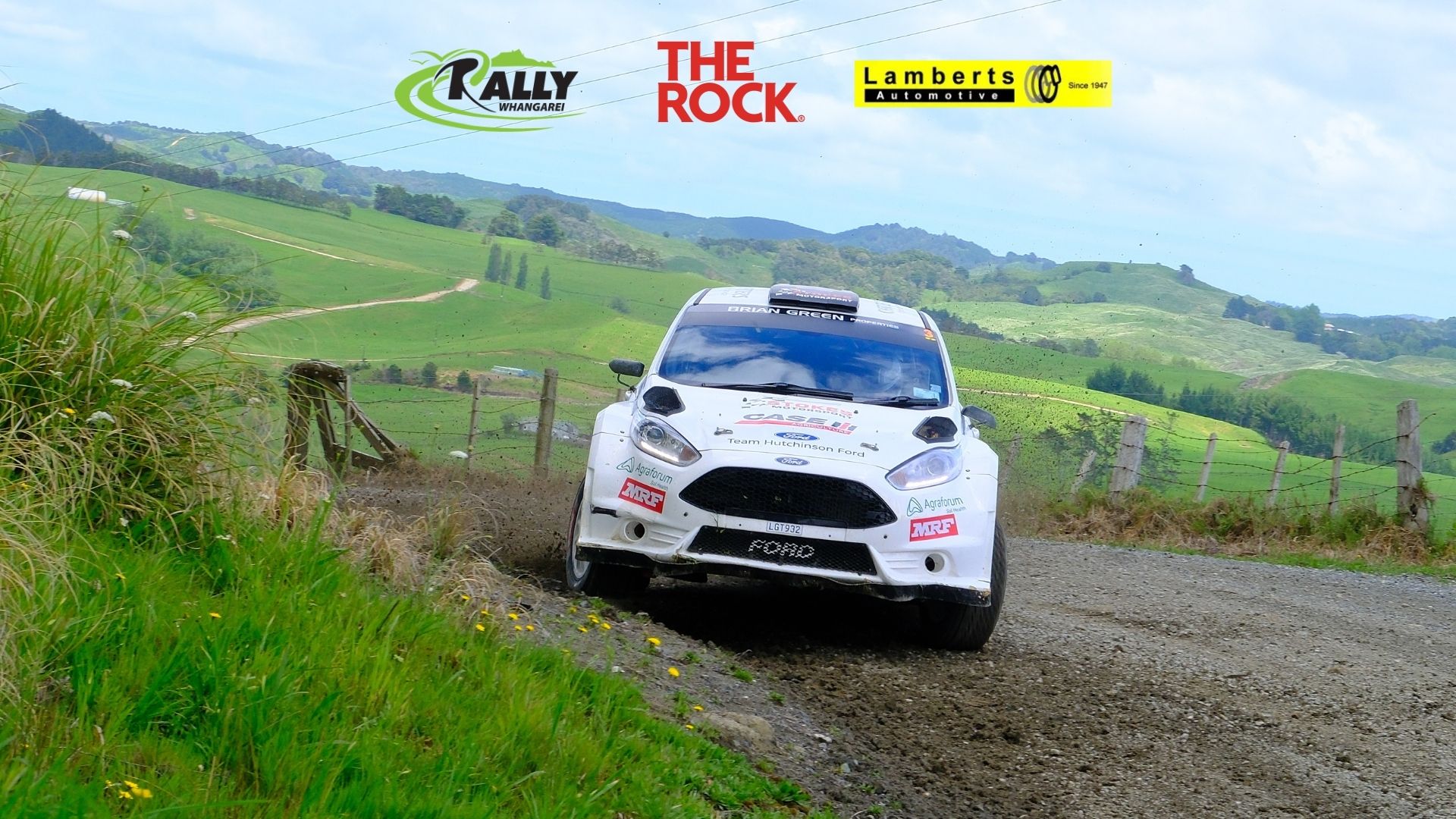 A white rally car speeds along a dirt road, kicking up dust, with green rolling hills, a fence, and event sponsor logos visible in the background and top of the image.