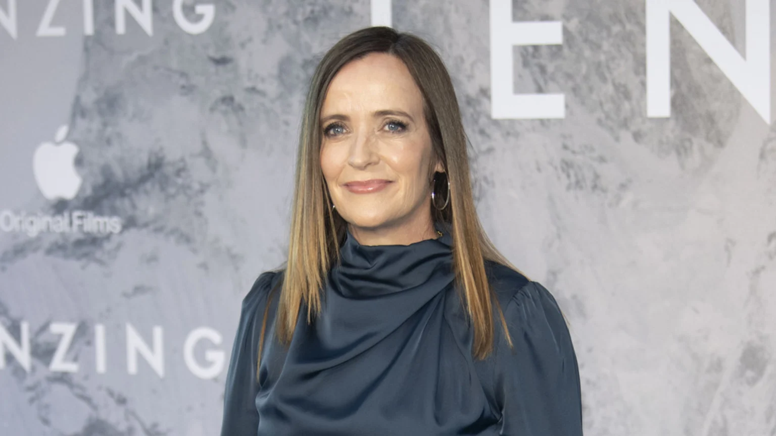 Jennifer Peedom smiles, wearing a dark blue satin top and hoop earrings, standing in front of an "Apple Original Films" backdrop with "TENZING" text.