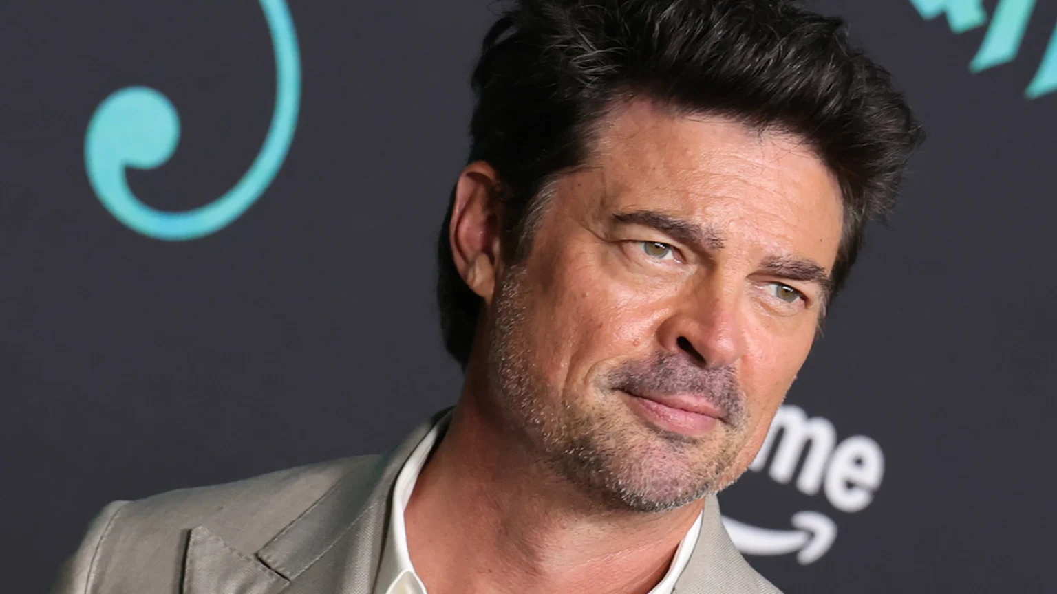 Actor Karl Urban is seen in a close-up, his dark hair and stubble framing his face as he looks slightly right with a faint smile, wearing a light jacket against a dark background.