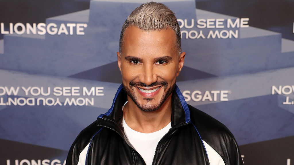 A close-up portrait of a man with slicked-back blonde hair and beard, smiling broadly in a black and blue jacket over a white t-shirt. He stands before a blue and grey geometric pattern backdrop featuring mirrored text related to Lionsgate and the film Now You See Me.