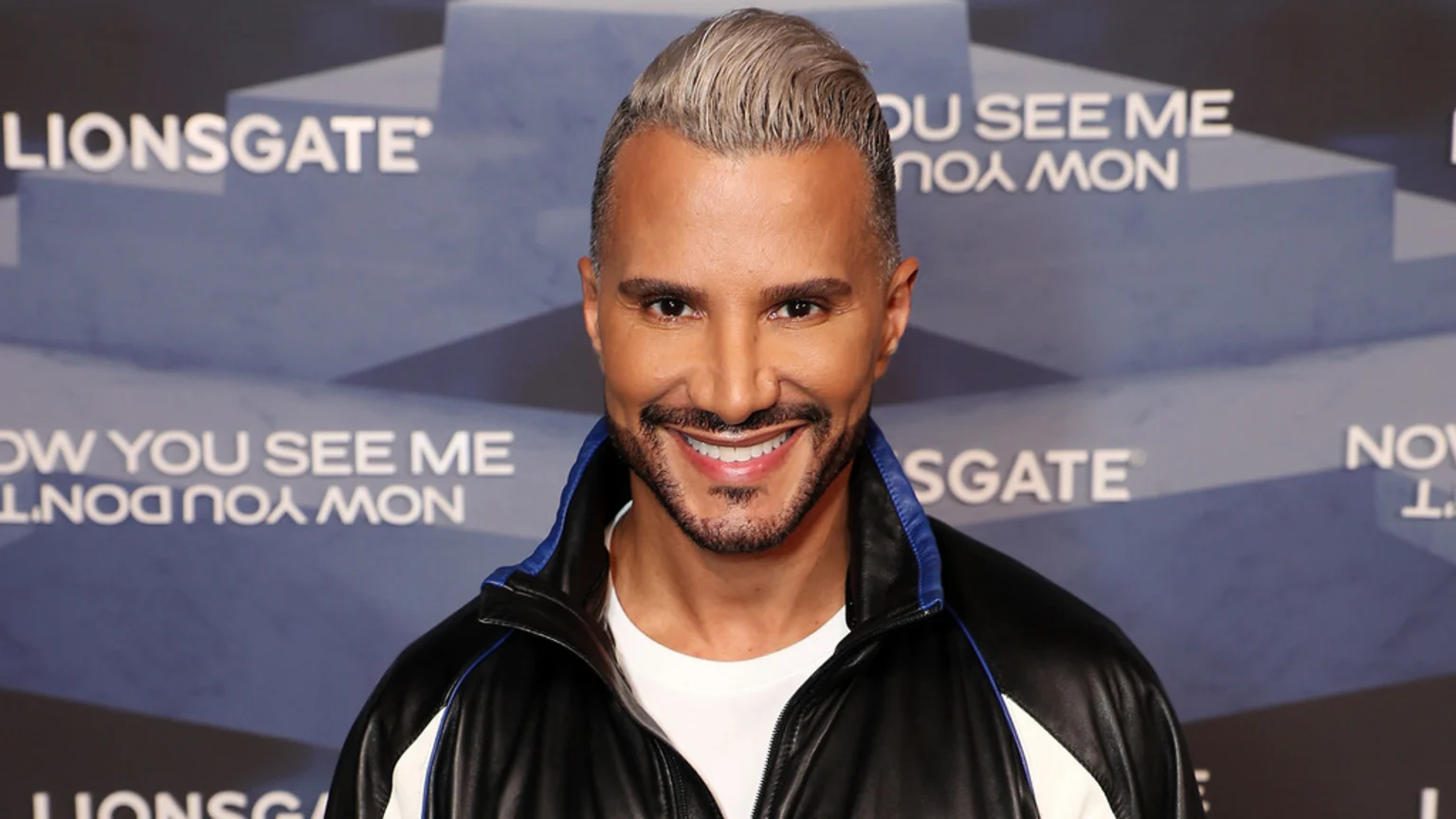 A close-up portrait of a man with slicked-back blonde hair and beard, smiling broadly in a black and blue jacket over a white t-shirt. He stands before a blue and grey geometric pattern backdrop featuring mirrored text related to Lionsgate and the film Now You See Me.