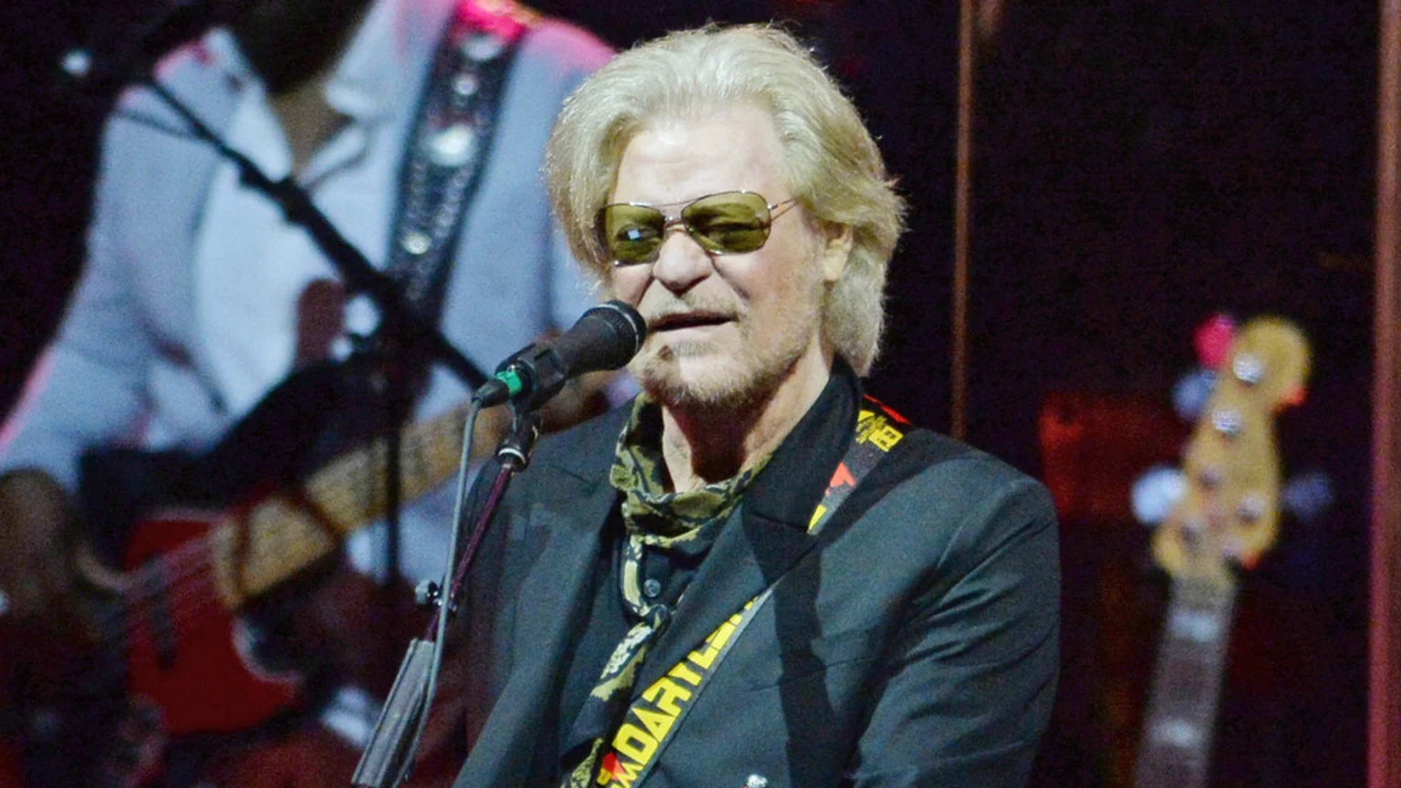 Daryl Hall, a man with blonde hair and a beard, wearing sunglasses, a dark suit, and a patterned scarf, sings into a microphone with a guitar strap over his shoulder on stage.