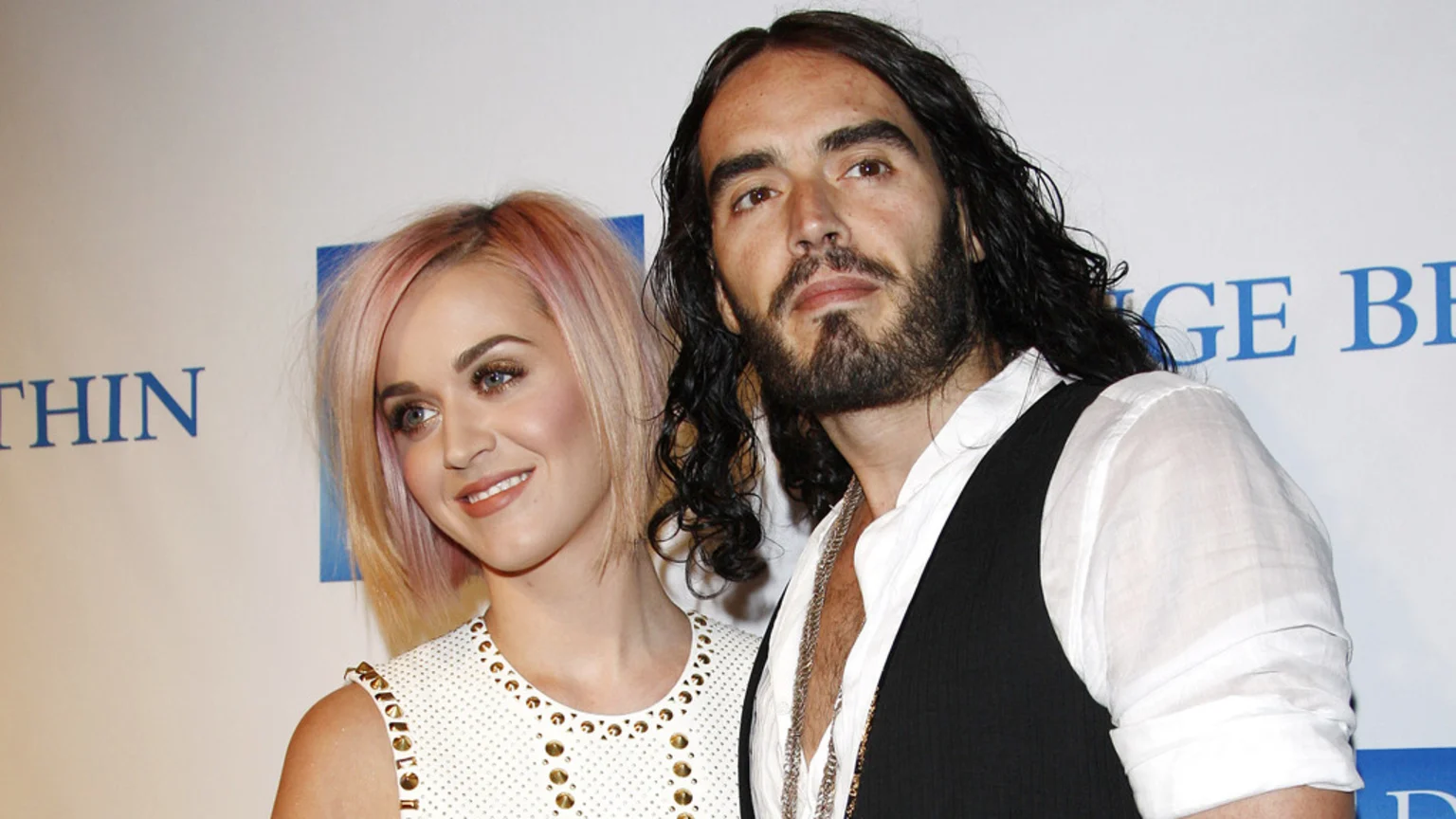Katy Perry, smiling with pinkish-blonde hair, stands beside a serious-looking Russell Brand, who has long dark hair and a beard, against a white and blue background.