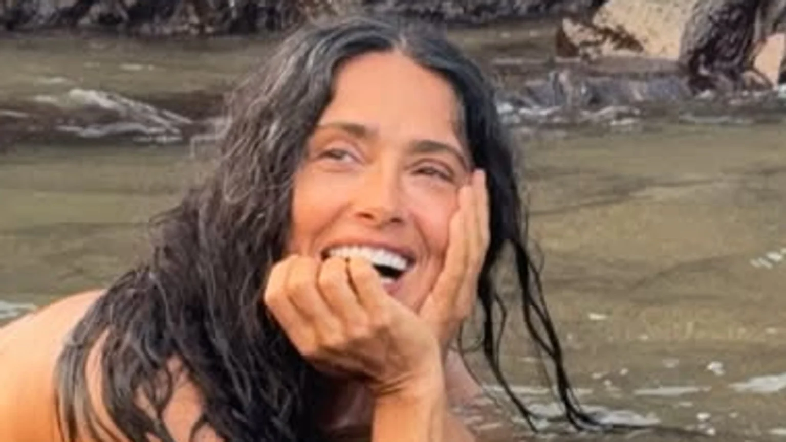 A joyful woman with wet, dark, wavy hair and visible grey strands smiles broadly, resting her hand on her chin while partially submerged in natural water with rocks in the background.