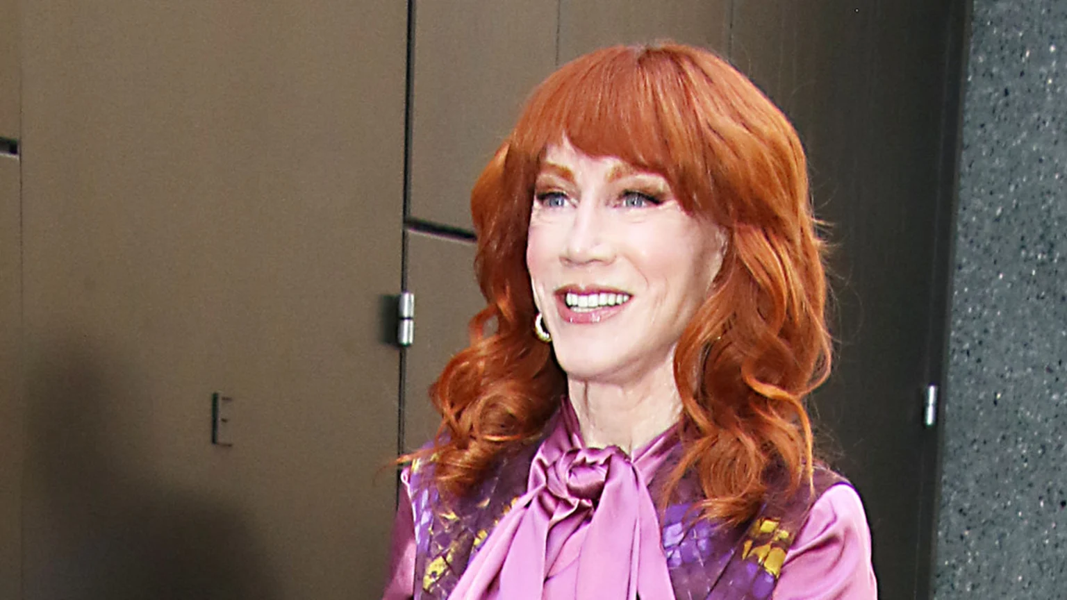 Kathy Griffin smiles, showing her teeth, with vibrant red hair, wearing a pink satin blouse with a bow tie and a purple patterned top.